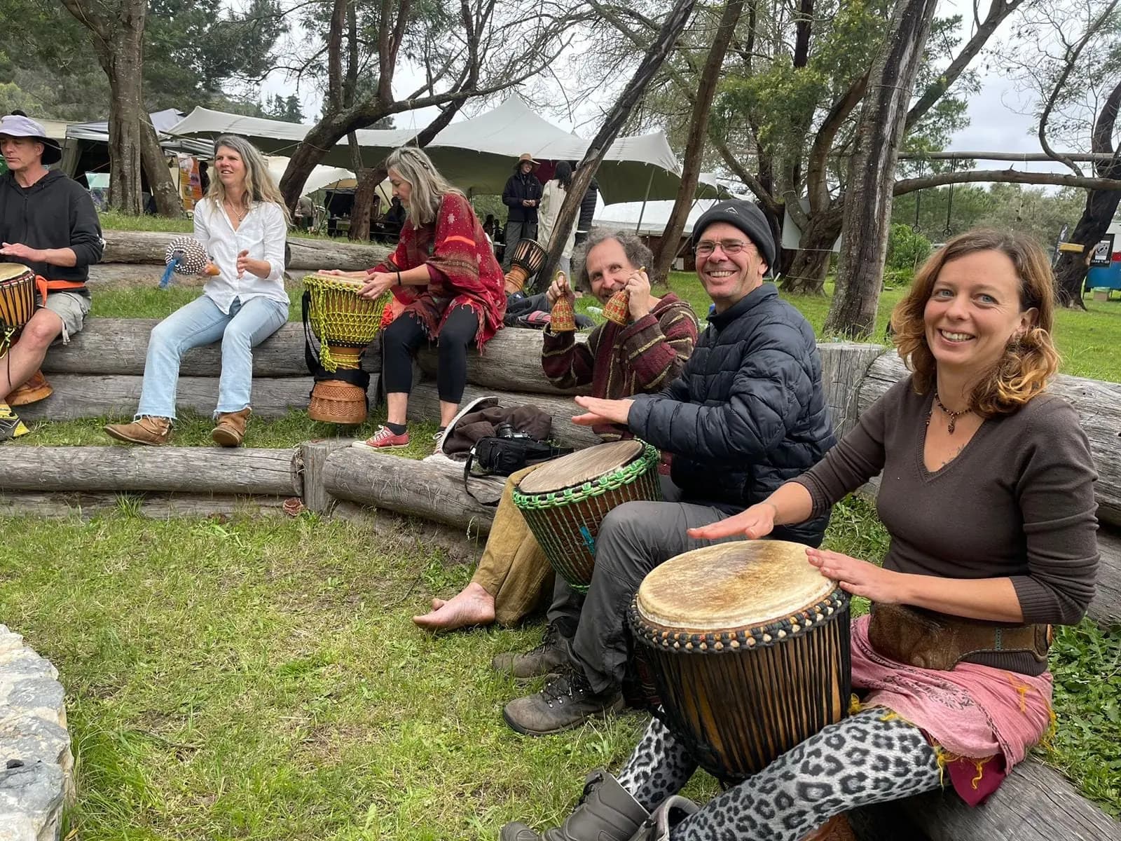 A group plays djembe drums together seated on log benches under the trees