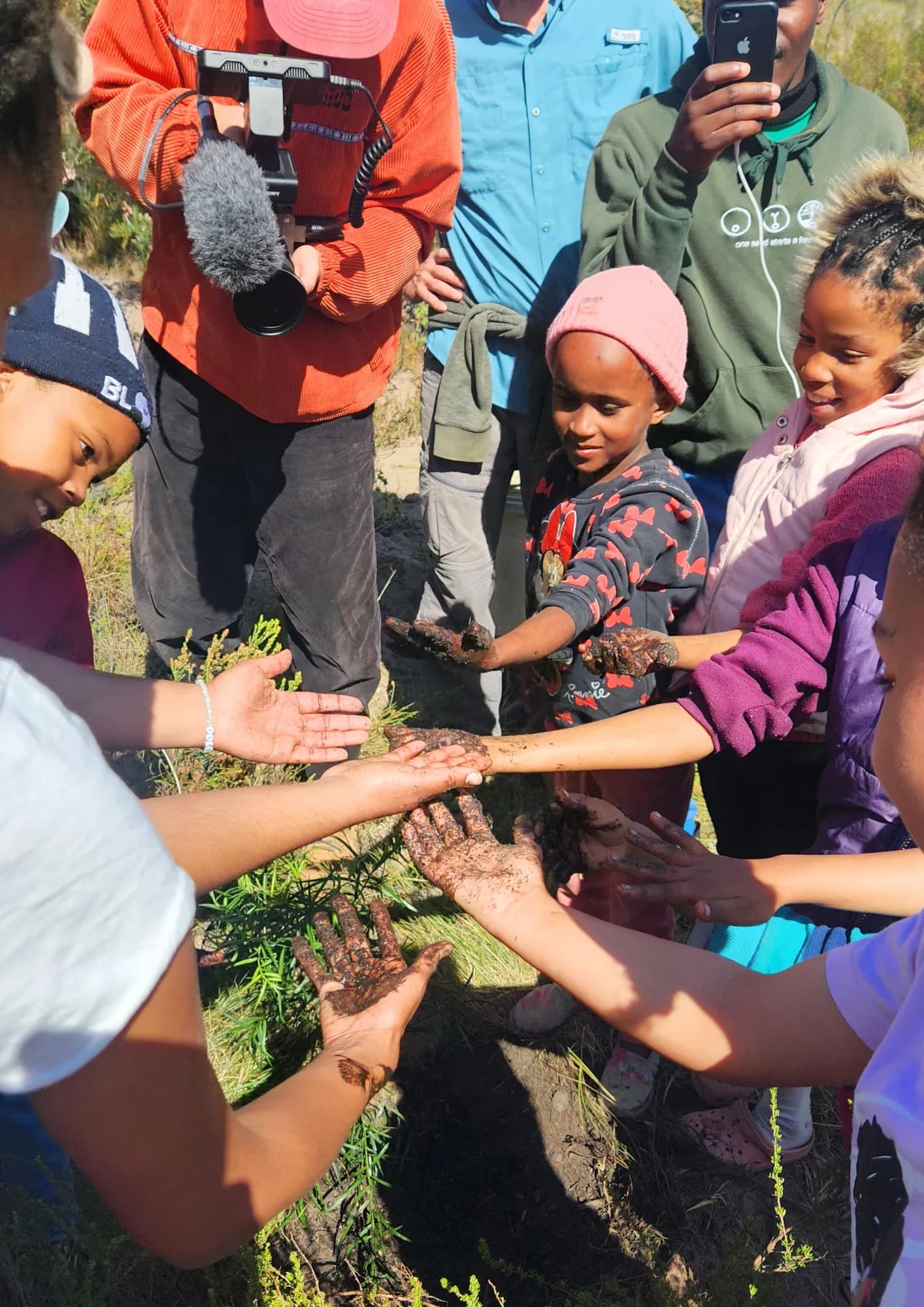 A group of children and adults hold out mud-covered hands together after digging, with a camera crew filming nearby